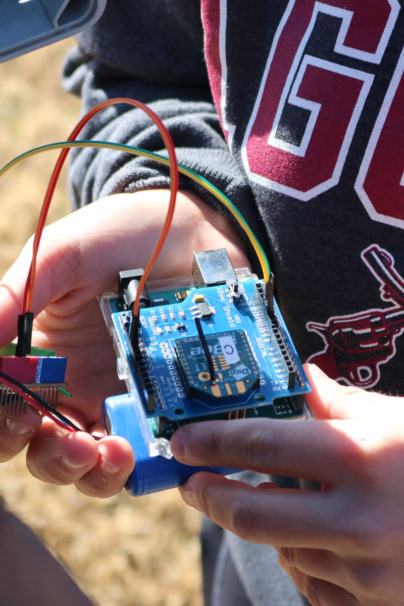 Hands holding a small Arduino-style circuit board with jumper wires connected, outdoors.